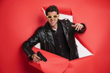 Siyah deri ceket giyen beyaz bir erkek portresi, silah taşıyor, aksiyon gerilim kitabı kapağı tasarımı için ideal yırtık bir kağıt deliğinden dışarı bakıyor.