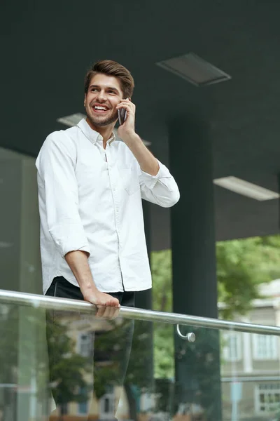 ヨーロッパのビジネスマンが携帯電話で話してどこかを探してください 若い男が市内の近代的なオフィスビルのバルコニーに立っている 髭を生やした男が白いシャツとジーンズを着ている — ストック写真