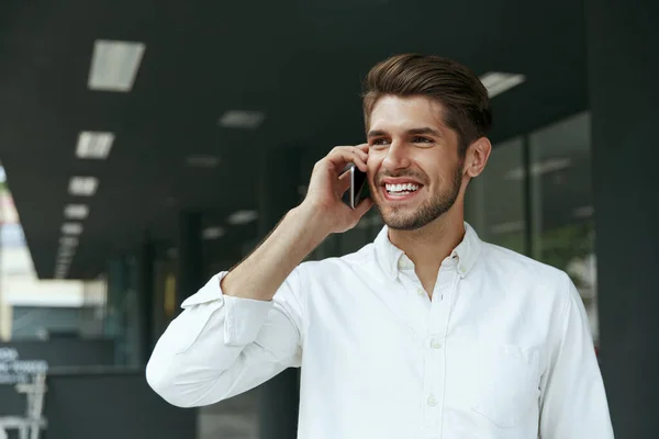 ヨーロッパのビジネスマンが携帯電話で話してくれました 若い男は市内の近代的なオフィスビルの背景に立っている 髭を生やしたスタイリッシュな男が白いシャツを着ている — ストック写真