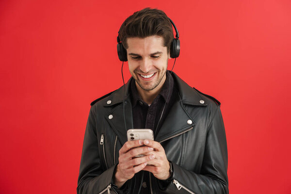 Young unshaven man smiling while using cellphone and headphones isolated over red wall