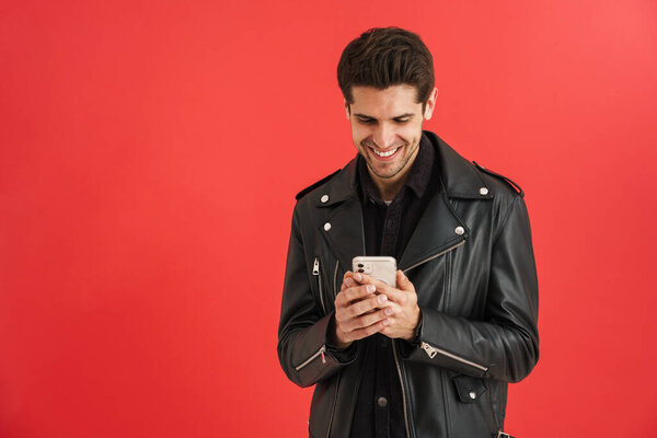 Young unshaven man smiling while using mobile phone isolated over red wall