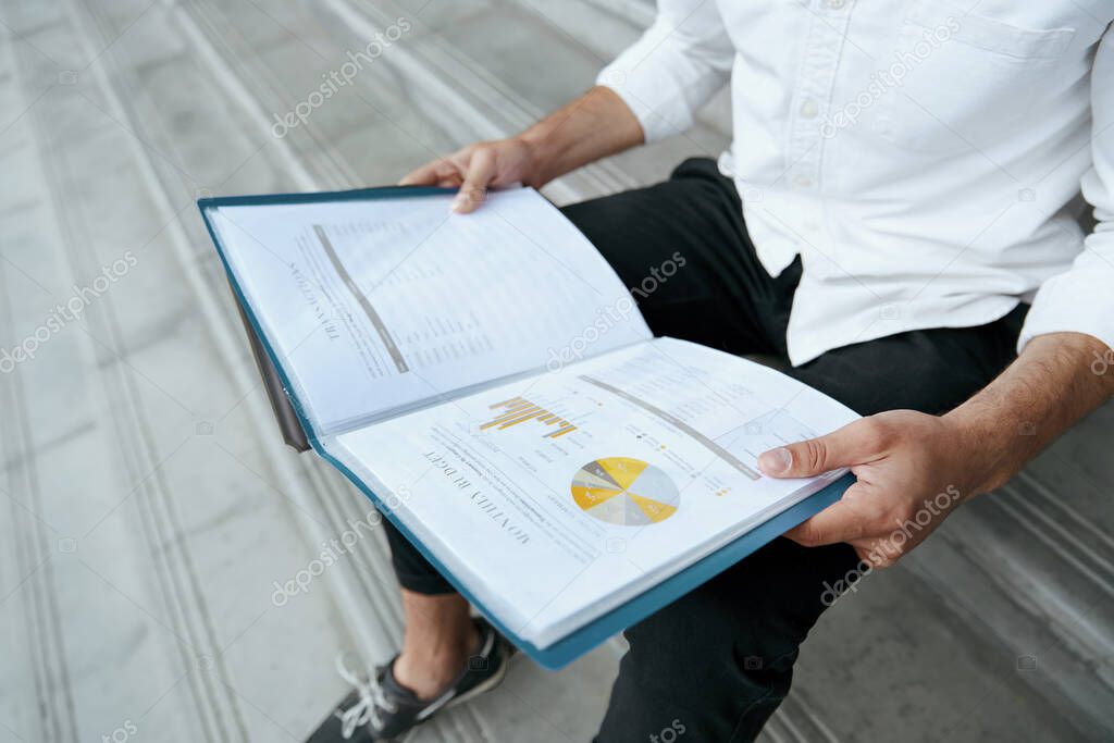Businessman sitting on stairs and watching open documents of monthly budget with chart pie. Close up view. Stylish guy wear white shirt and black jeans. Daytime