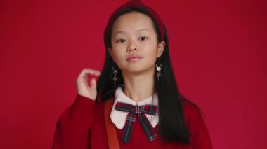 Kendine güvenen küçük Asyalı kız kırmızı stüdyodaki kamerada görüntüsünü düzeltiyor.