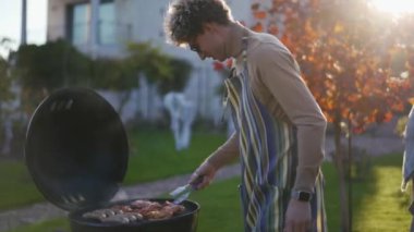 Gülümseyen kıvırcık saçlı adam piknikte ızgara sosis hazırla.