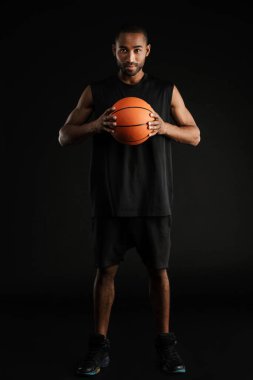 Genç sporcu siyah arka planda basketbolla poz verirken kameraya bakıyor.