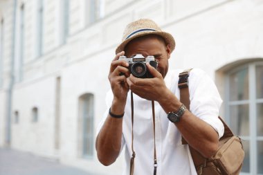 Siyahi adam Avrupa 'da dijital kamerada fotoğraf çekiyor. Genç adam hasır şapka, beyaz gömlek ve kol saati takıyor. Kamera çantası tutan kişi. Seyahat kavramı. Serbest çalışmanın bir fikri. Güneşli gündüz