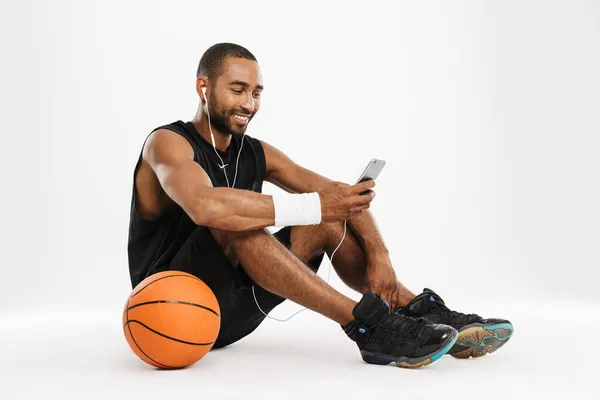 幸せなアフリカの若いバスケットボール選手は 白い背景に隔離されたイヤフォンや携帯電話で音楽を聴いて座って — ストック写真