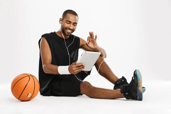 幸せなアフリカの若いバスケットボール選手は白い背景に隔離されたタブレットコンピュータやイヤフォンを介して話して座っている — ストック写真