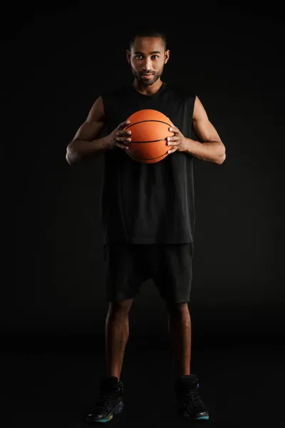 年轻的运动员一边拿着黑色背景的篮球 一边看着镜头 — 图库照片