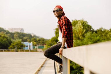 Genç siyahi adam dışarıda çitin üzerinde otururken gülümsüyor.