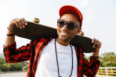 Sıradan giyinmiş gülümseyen Afrikalı genç adam dışarıda kaykay tutuyor.