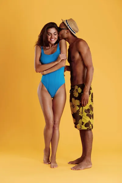 Black man kissing his pleased girlfriend. Young beautiful romantic couple. Guy and european woman wear beach clothes. Concept of summer beach vacation. Isolated on yellow background. Studio shoot