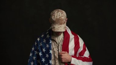 Amerikan bayrağının arkasına saklanıp siyah stüdyodaki kameraya bakan ciddi bir asker.