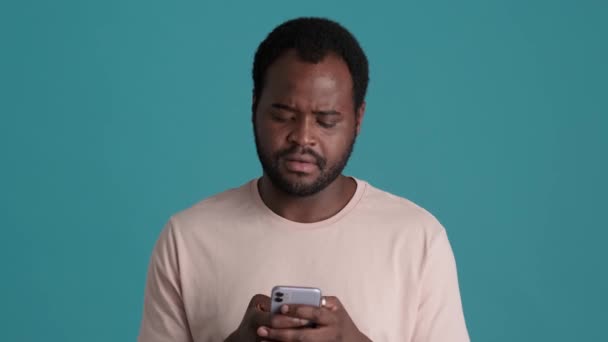 青のスタジオで電話でTシャツの思考とテキストメッセージを身に着けているアフリカ人男性 — ストック動画