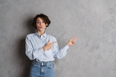 Genç esmer kadın şaşkınlığını dile getiriyor ve parmaklarını gri duvarın üzerinde izole bir şekilde gösteriyor.
