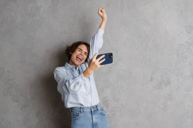 Gri duvarda izole edilmiş cep telefonuyla çevrimiçi oyun oynarken el kol hareketi yapan genç bir kadın.