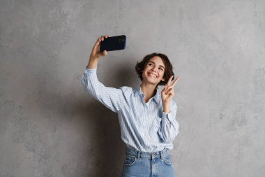Genç esmer kadın, gri duvardan izole edilmiş cep telefonuyla selfie çekerken el kol hareketi yapıyor.
