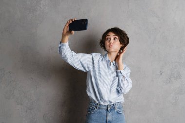 Genç esmer kadın, gri duvardan izole edilmiş cep telefonuyla selfie çekerken yüzünü buruşturuyor.