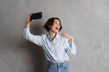 Gri duvardan izole edilmiş cep telefonuyla selfie çeken esmer bir kadın.