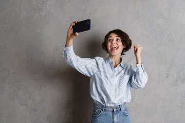 Genç esmer kadın, gri duvardan izole edilmiş cep telefonuyla selfie çekerken el kol hareketi yapıyor.