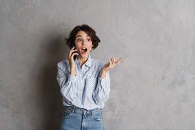 Genç esmer kadın, gri duvarın üzerinde cep telefonuyla konuşurken şaşkınlığını dile getiriyor.
