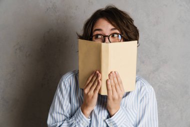 Gözlük takan genç bir kadın gri duvarın üzerinde kitap ile poz verirken yana bakıyor.