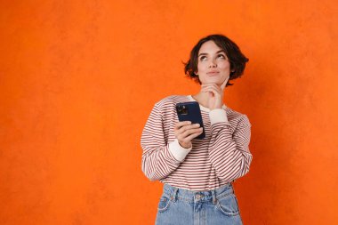 Çizgili tişörtlü, turuncu duvarda izole edilmiş cep telefonu kullanan esmer bir kadın.