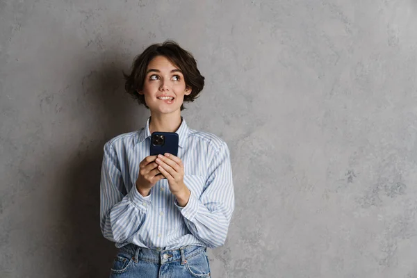 Молодая Брюнетка Кусает Губу Использует Мобильный Телефон Изолированный Над Серой — стоковое фото