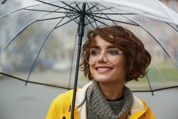 Young girl in yellow raincoat with transparent umbrella in city. Young beautiful woman wearing rain outfit and eyeglasses while walking on street. Concept of modern woman lifestyle at autumn.