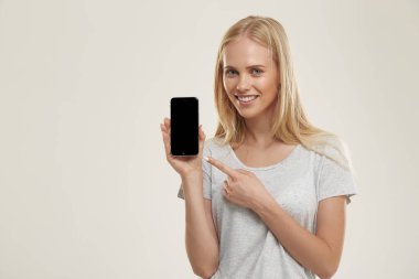 Genç gülümseyen Avrupalı kız cep telefonunu tutuyor ve gösteriyor. Mavi gözlü güzel sarışın genç kız beyaz tişört giyer ve kameraya bakar. Beyaz arka planda izole edilmiş. Stüdyo çekimi. Boşluğu kopyala