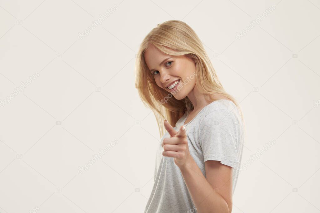 Young smiling european girl point with finger to camera. Pretty blonde female teenager with blue eyes wear white t-shirt and looking at camera. Isolated on white background. Studio shoot. Copy space