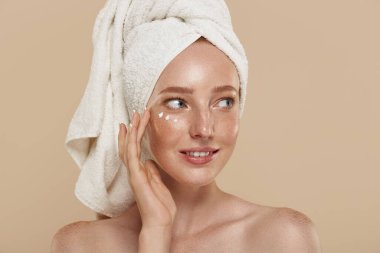 Çıplak omuzları, göz altı bantları ve kafasında havlu olan beyaz bir kız. Çilli genç ve güzel bir kadının kısmi görüntüsü. Doğal kadın güzelliği. Bej arkaplanda izole edilmiş.