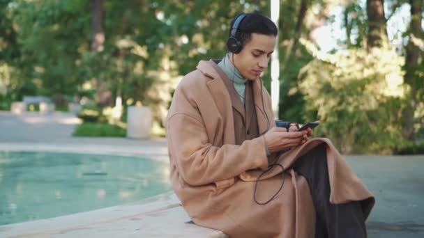 公园里的阿拉伯男子用耳机用电话发短信的侧视图 — 图库视频影像