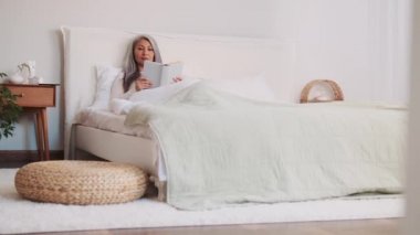 Neşeli Asyalı kadın kitap okuyor ve ondan sonra yataktaki telefona bakıyor.