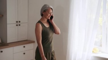 Pozitif Asyalı kadın telefonla konuşuyor ve mutfaktaki pencereye bakıyor.