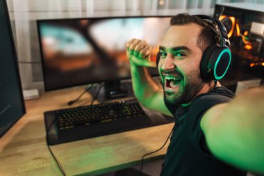 Mutlu genç adam evde bilgisayar başında video oyunu oynarken fotoğraf çekiyor ve başarıyı kutluyor.