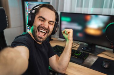 Mutlu genç adam evde bilgisayar başında video oyunu oynarken fotoğraf çekiyor ve başarıyı kutluyor.