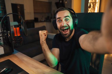 Mutlu genç adam evde bilgisayar başında video oyunu oynarken fotoğraf çekiyor ve başarıyı kutluyor.