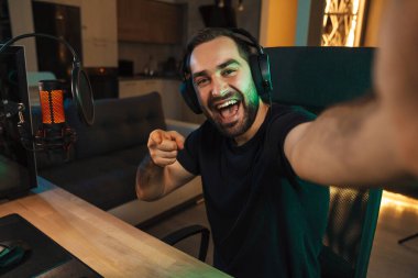 Mutlu genç adam evde bilgisayar başında video oyunu oynarken fotoğraf çekiyor ve başarıyı kutluyor.