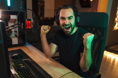 Mutlu genç oyuncu kulaklıkla bilgisayar başında oyuncularla sohbet ederek oyun yarışmasında zaferi kutluyor.