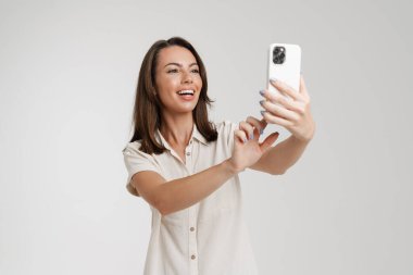 Genç Avrupalı kadın cep telefonuyla selfie çekiyor. Beyaz arka planda izole edilmiş.