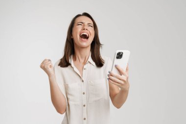 Genç bir kadın başarılı bir jest yapıyor ve beyaz arka planda izole edilmiş cep telefonu kullanıyor.