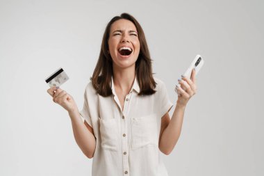 Beyaz arka planda izole edilmiş cep telefonu ve kredi kartıyla poz veren heyecanlı genç kadın.