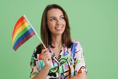 Genç Avrupalı kadın yeşil arka planda izole edilmiş gökkuşağı bayrağıyla poz verirken gülümsüyor