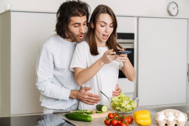 Evde birlikte yemek pişirirken cep telefonu kullanan ve sarılan çok ırklı genç bir çift.