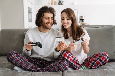 Genç mutlu çift ellerinde kredi kartı ve joystickle evdeki kanepede oturuyorlar.