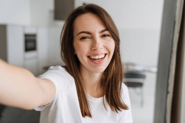 Salondaki gülümseyen genç kadın evde dikilip selfie çekiyor.