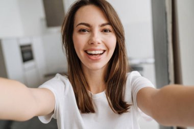Salondaki gülümseyen genç kadın evde dikilip selfie çekiyor.