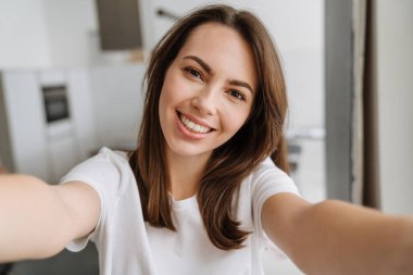 Salondaki gülümseyen genç kadın evde dikilip selfie çekiyor.