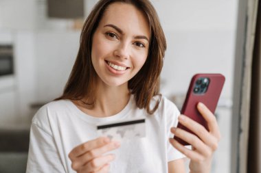 Genç beyaz kadın içeride kredi kartı ve cep telefonuyla poz verirken gülümsüyor.